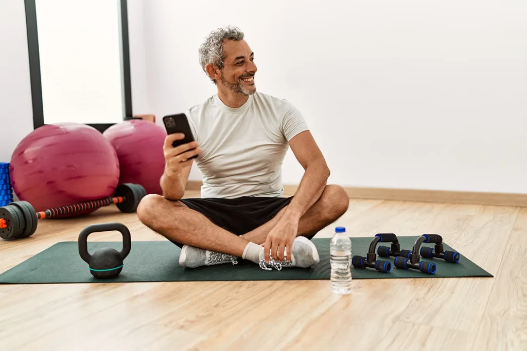 Happy, healthy, active older age grey-haired man sitting on yoga mat with weights next to hime in a gym environment, looking at fitness data on smartphone app connected to his hearing aids with health tracking 